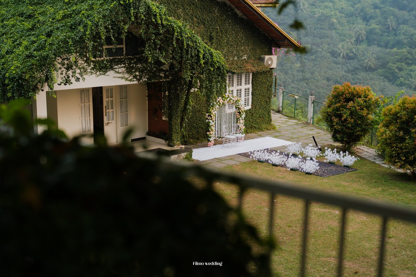 A real wedding setup at Naz's Farm House: floral arch and aisle beside the ivy-covered house, with misty forested hills behind